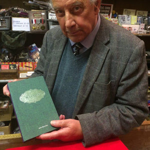 Roy Walton behind the counter in Tam Shepherds Trick Shop, holding a copy of his book 'The Complete Walton: Volume 3'. 
